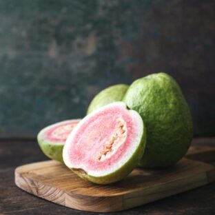 4 guavas (2 whole and 2 cut to reveal pink flesh and seeds) placed on a wooden cutting board, matte black background