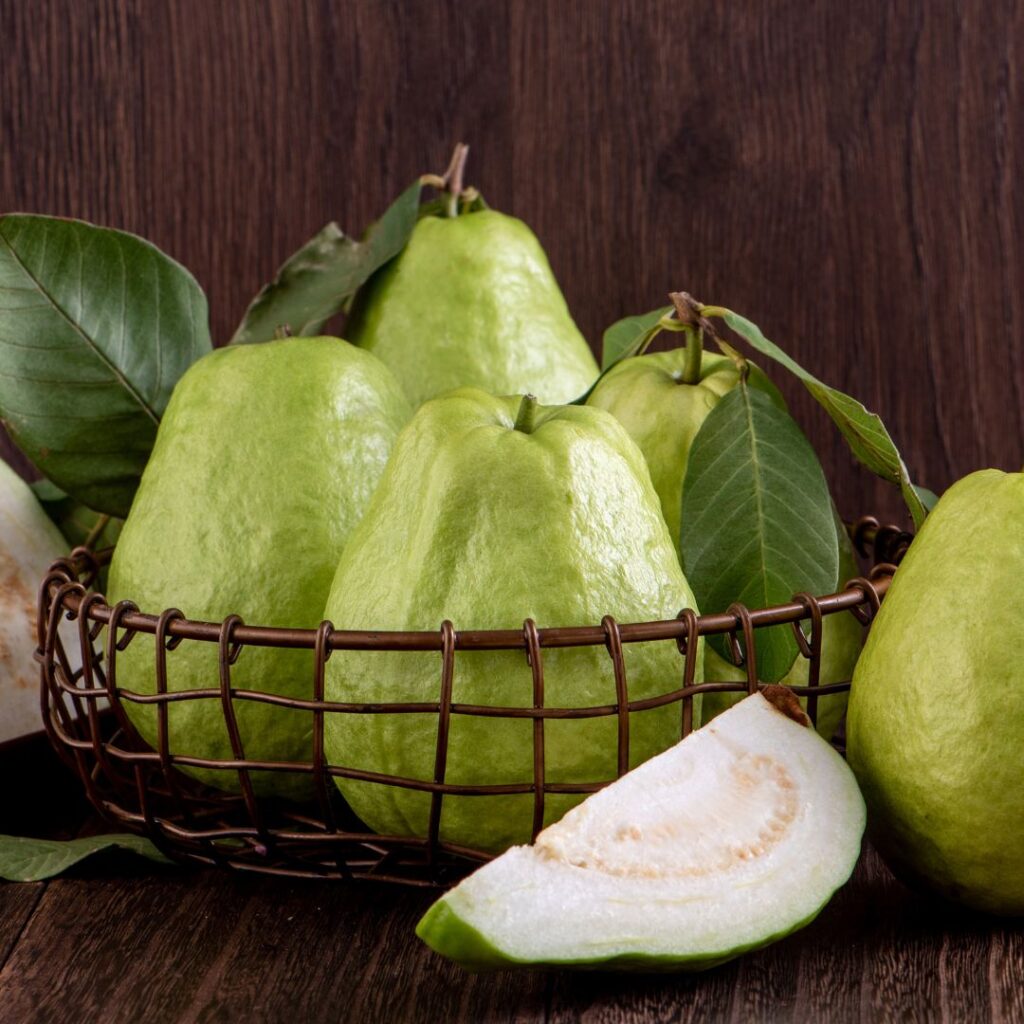 4 guavas in an iron basket (black square mesh style), 1/4 of a white-fleshed guava is cut and placed in front of the basket containing 4 guavas, 1 guava is placed to the right of the cut guava.